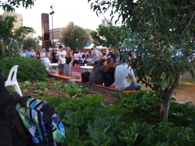 Three people are sitting with their backs to the camera on the edge of a waist-high garden bed. The garden bed is full of vegetables and has a fruit tree on the right hand side of the photo. Beyond are crowds of people walking or gaterhing around little tables, and in the farthest distance of the photo, about twenty metres away, are white tents housing market stalls, mostly hidden by the crowds in front of them.